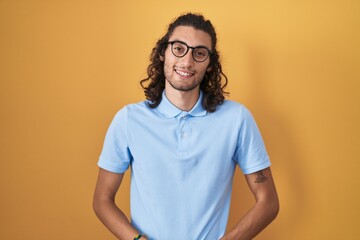 Young hispanic man standing over yellow background with hands together and crossed fingers smiling relaxed and cheerful. success and optimistic