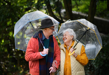 Happy senior couple walking with umbrellas in city park together.