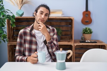 Young hispanic man drinking coffee from french coffee maker serious face thinking about question with hand on chin, thoughtful about confusing idea
