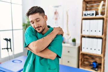 Fototapeta premium Young physiotherapist man working at pain recovery clinic hugging oneself happy and positive, smiling confident. self love and self care