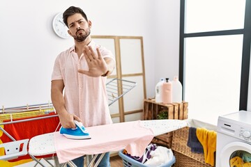 Young man with beard ironing clothes at home doing stop sing with palm of the hand. warning expression with negative and serious gesture on the face.