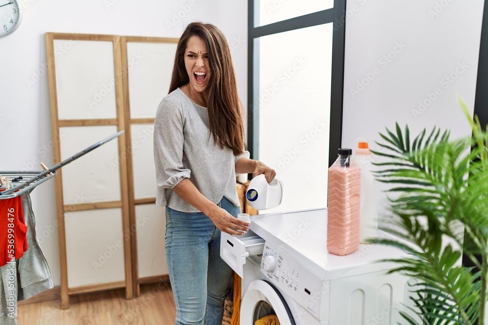 Poster young brunette woman putting detergent in washing machine angry and mad screaming frustrated and fur