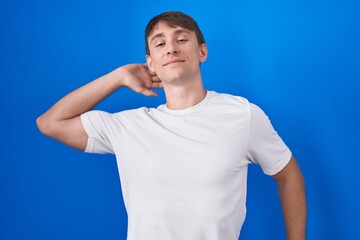 Caucasian blond man standing over blue background stretching back, tired and relaxed, sleepy and yawning for early morning