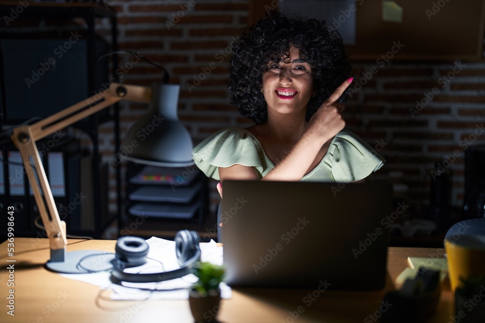 Poster young brunette woman with curly hair working at the office at night with a big smile on face, pointi