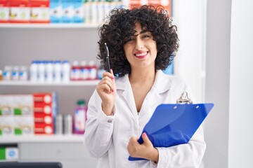 Young middle east woman pharmacist smiling confident holding clipboard at pharmacy