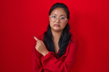 Asian young woman standing over red background pointing aside worried and nervous with forefinger,...