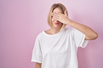 Young caucasian woman standing over pink background covering eyes with hand, looking serious and sad. sightless, hiding and rejection concept