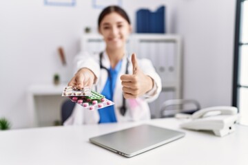Young hispanic doctor woman wearing doctor uniform holding prescription pills at clinic smiling happy and positive, thumb up doing excellent and approval sign