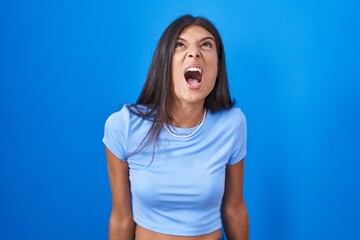 Fototapeta na wymiar Brunette young woman standing over blue background angry and mad screaming frustrated and furious, shouting with anger. rage and aggressive concept.