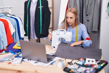 Young blonde woman tailor using sewing machine and laptop at tailor shop