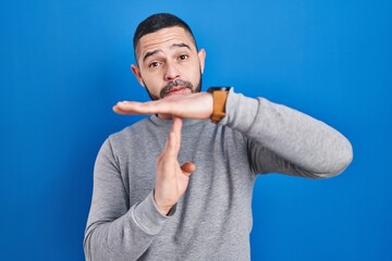 Hispanic man standing over blue background doing time out gesture with hands, frustrated and...