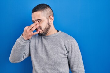 Hispanic man standing over blue background smelling something stinky and disgusting, intolerable smell, holding breath with fingers on nose. bad smell