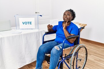 Young african woman sitting on wheelchair voting putting envelop in ballot box thinking...