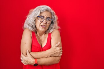 Middle age woman with grey hair standing over red background shaking and freezing for winter cold with sad and shock expression on face