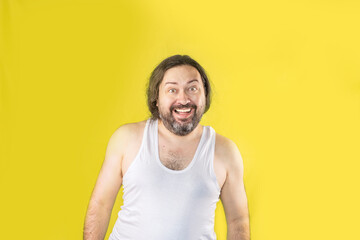 Obraz premium A joyful, unshaven man in a white undershirt stands and smiles exaggeratedly. Unemployed slovenly dressed man from Eastern Europe.