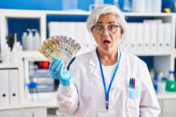 Middle age woman with grey hair working at scientist laboratory holding polish zloty banknotes scared and amazed with open mouth for surprise, disbelief face
