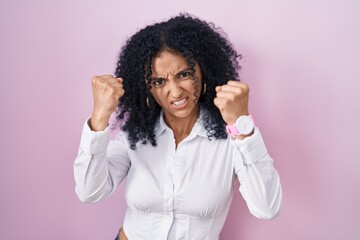 Hispanic woman with curly hair standing over pink background angry and mad raising fists frustrated and furious while shouting with anger. rage and aggressive concept.