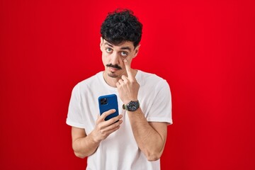 Hispanic man using smartphone over red background pointing to the eye watching you gesture, suspicious expression