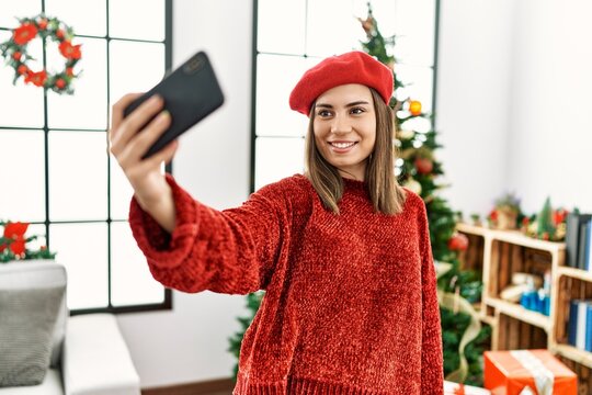 Young hispanic girl making selfie by the smartphone standing by christmas tree at home.