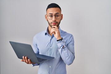 Young hispanic man working using computer laptop thinking concentrated about doubt with finger on chin and looking up wondering