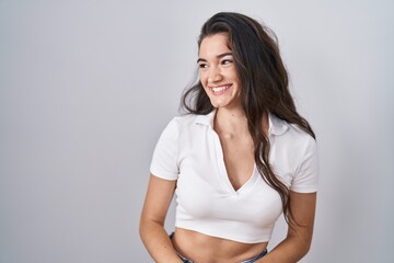 Young teenager girl standing over white background looking away to side with smile on face, natural expression. laughing confident.