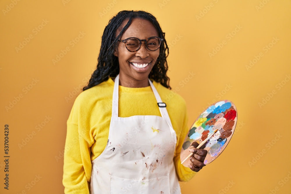 Poster african woman holding painter palette winking looking at the camera with sexy expression, cheerful a