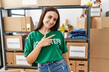 Young brunette woman wearing volunteer t shirt at donations stand cheerful with a smile on face pointing with hand and finger up to the side with happy and natural expression