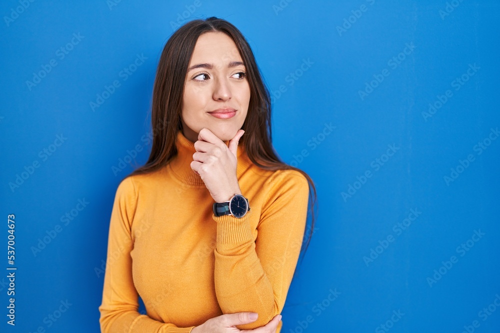 Poster young brunette woman standing over blue background looking confident at the camera smiling with cros