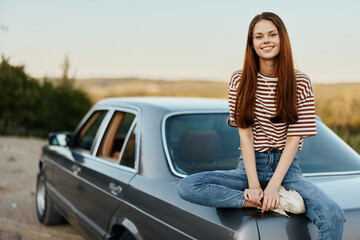 A young woman sits on the trunk of a car and rests after a difficult road and admires nature with a beautiful view. Stopping is also part of the journey