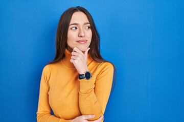 Young brunette woman standing over blue background looking confident at the camera smiling with crossed arms and hand raised on chin. thinking positive.
