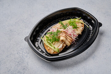 Bruschetta with smoked eel, young cheese and cucumber in black plastic container, close up. Designer food concept for take away or delivery