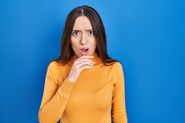 Young brunette woman standing over blue background looking fascinated with disbelief, surprise and amazed expression with hands on chin