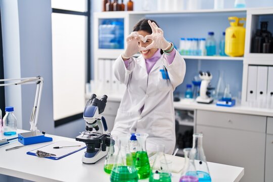 Young brunette woman working at scientist laboratory smiling in love showing heart symbol and shape with hands. romantic concept.