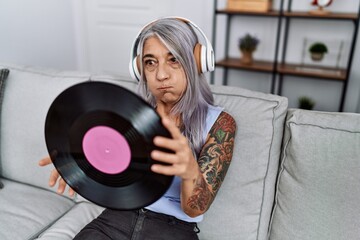 Middle age grey-haired woman listening to music holding vinyl disc at home puffing cheeks with funny face. mouth inflated with air, catching air.