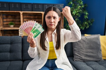 Young latin woman holding norwegian money annoyed and frustrated shouting with anger, yelling crazy...