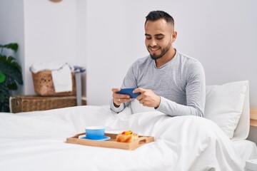 Young hispanic man having breakfast playing video game at bedroom