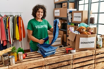 Young hispanic woman wearing volunteer uniform folding jeans at charity center