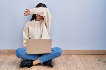 Young woman using laptop sitting on the floor at home covering eyes with arm, looking serious and...