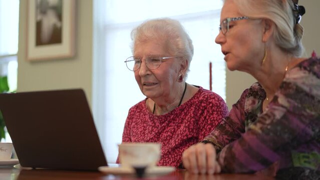 Push in to happy elderly middle aged woman sitting at table with grown up daughter, watching family or journey photos on computer. Smiling blonde lady listening to older, using laptop.