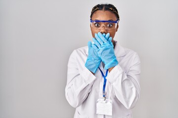 African american woman with braids wearing scientist robe shocked covering mouth with hands for mistake. secret concept.
