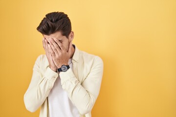 Young hispanic man standing over yellow background with sad expression covering face with hands while crying. depression concept.