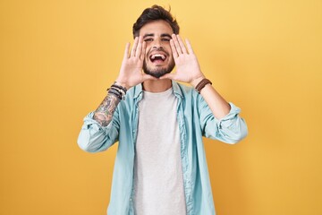Young hispanic man with tattoos standing over yellow background smiling cheerful playing peek a boo...