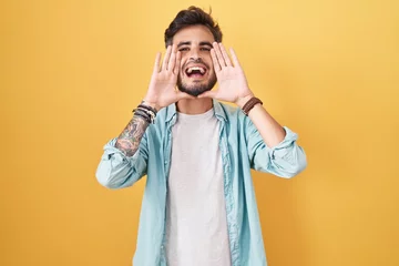 Fotobehang Young hispanic man with tattoos standing over yellow background smiling cheerful playing peek a boo with hands showing face. surprised and exited © Krakenimages.com