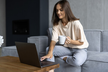 Fototapeta premium Beautiful young woman working on laptop while sitting at the living room, drinking coffee