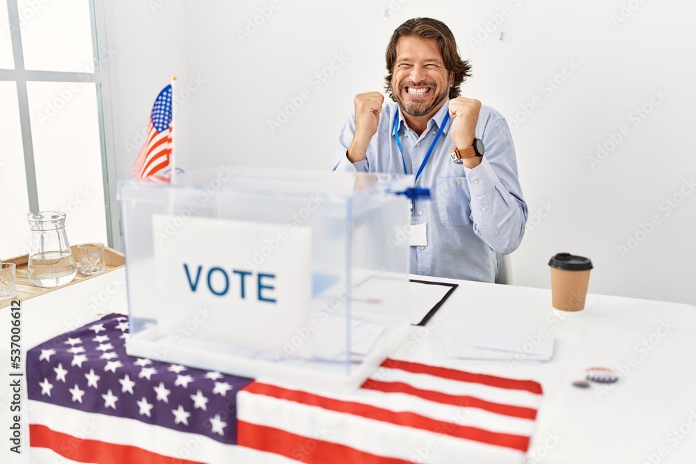 Wall mural Handsome middle age man sitting at voting stand excited for success with arms raised and eyes closed celebrating victory smiling. winner concept.