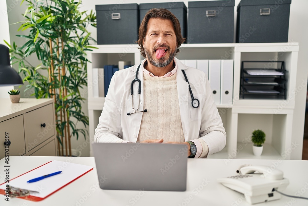 Sticker Handsome middle age doctor man working at the clinic sticking tongue out happy with funny expression. emotion concept.