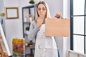 Young blonde painter woman holding wooden case at art studio covering mouth with hand, shocked and afraid for mistake. surprised expression