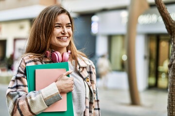 Young blonde woman wearing headphones smiling confident holding books at street