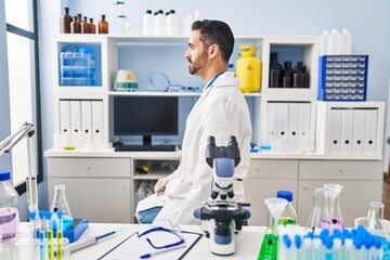 Young hispanic man with beard working at scientist laboratory looking to side, relax profile pose with natural face with confident smile.