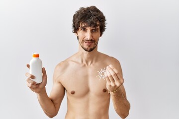 Fototapeta premium Young hispanic man standing shirtless holding sunscreen lotion doing money gesture with hands, asking for salary payment, millionaire business